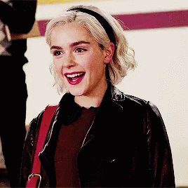 A Woman Wearing A Leather Jacket And A Headband Smiles GIF