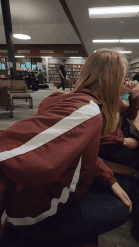 A Woman Wearing A Maroon Jacket With White Stripes On The Back Sits In A Library GIF