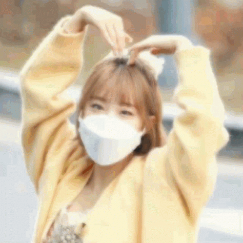 A Woman Wearing A Mask And A Yellow Sweater Is Making A Heart Shape With Her Hands . GIF