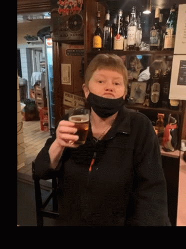 A Woman Wearing A Mask Holds A Glass Of Beer In Front Of A Sign That Says Public Telephone GIF