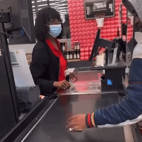 A Woman Wearing A Mask Is Standing At A Cash Register In Front Of A Sign That Says Coca Cola GIF