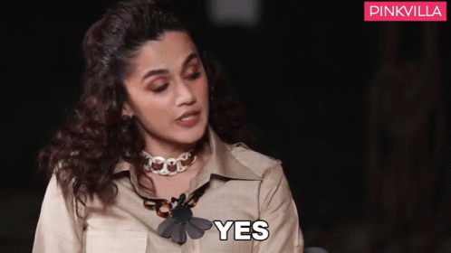 A Woman Wearing A Necklace And A Tan Shirt Is Sitting Down And Saying Yes . GIF