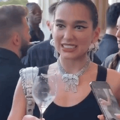 A Woman Wearing A Necklace And Earrings Is Holding A Wine Glass And A Cell Phone . GIF