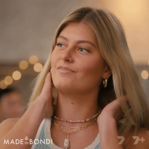 A Woman Wearing A Necklace And Earrings Is Smiling And Touching Her Hair GIF