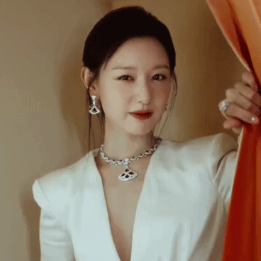 A Woman Wearing A Necklace And Earrings Is Standing In Front Of A Curtain GIF