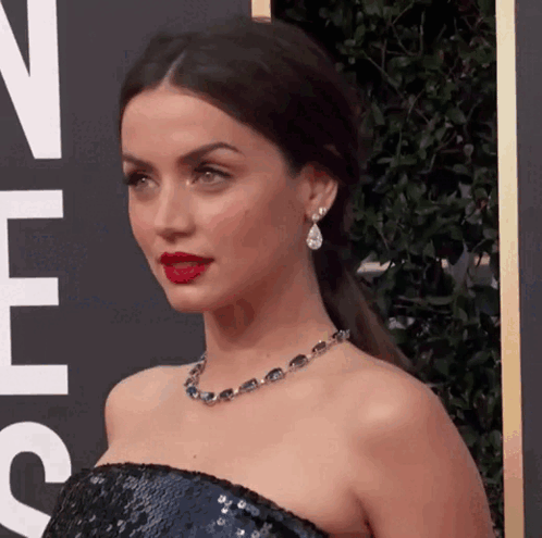 A Woman Wearing A Necklace And Earrings Is Standing In Front Of A Sign That Says ' N ' On It GIF