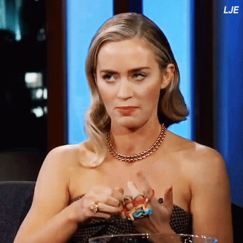 A Woman Wearing A Necklace And Rings Is Sitting In Front Of A Blue Wall With The Letters Lje On It GIF