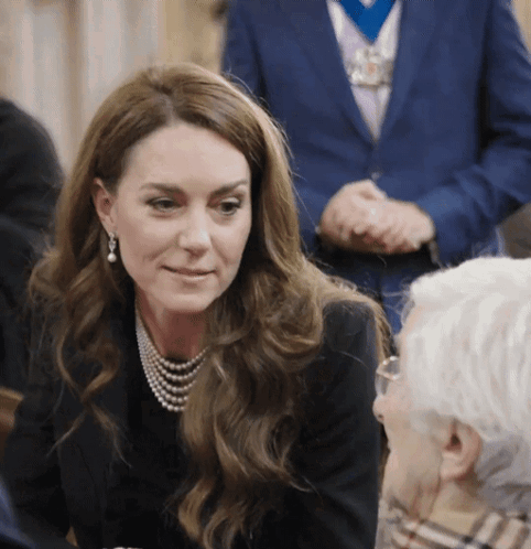 A Woman Wearing A Pearl Necklace And Earrings Talks To An Older Man GIF