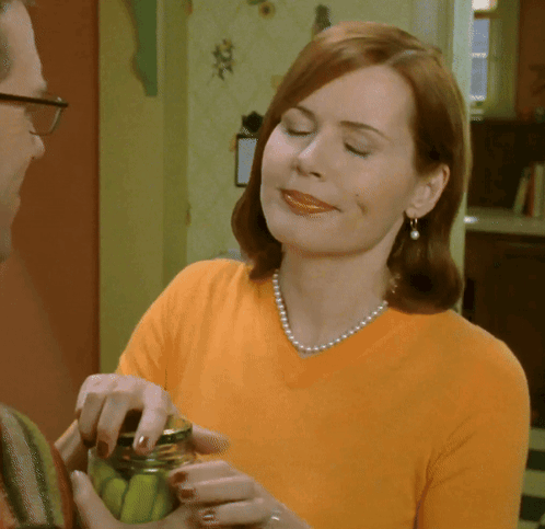 A Woman Wearing A Pearl Necklace Is Smiling While Holding A Jar Of Pickles GIF