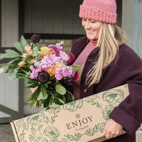 A Woman Wearing A Pink Hat Is Holding A Box Of Flowers That Says Enjoy Flowers GIF