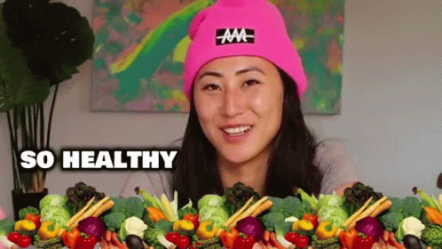 A Woman Wearing A Pink Hat Is Surrounded By Vegetables And The Words So Healthy Are Above Her . GIF