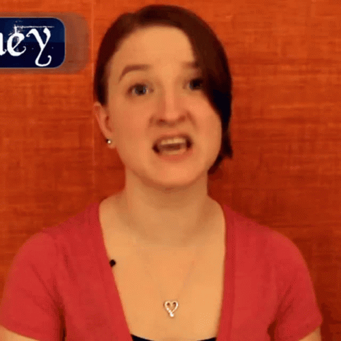 A Woman Wearing A Pink Shirt And A Heart Shaped Necklace Stands In Front Of A Sign That Says Hey GIF