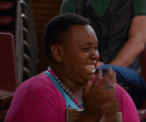 A Woman Wearing A Pink Sweater And A Blue Necklace Is Clapping Her Hands GIF
