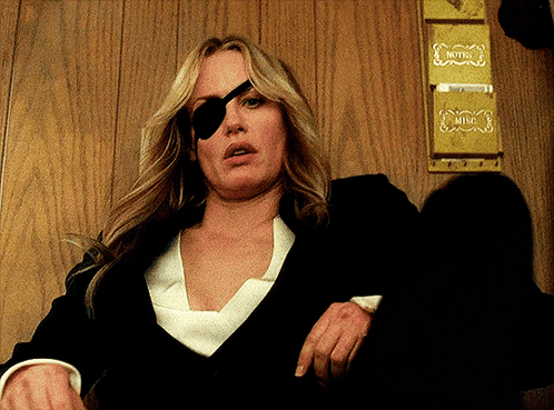 A Woman Wearing A Pirate Eye Patch Is Sitting In Front Of A Wall That Says Hotel GIF