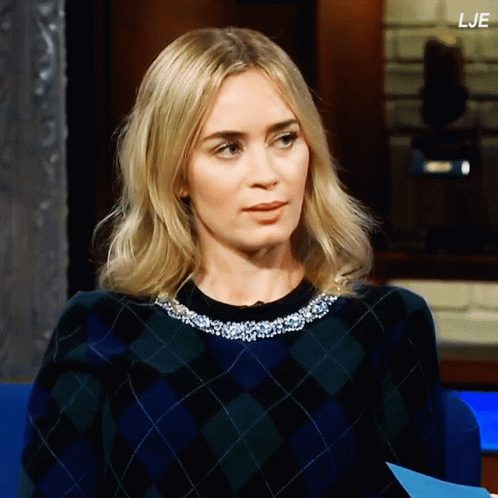 A Woman Wearing A Plaid Sweater Is Sitting In Front Of A Sign That Says " Lje " GIF
