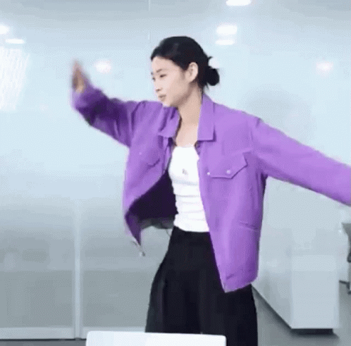 A Woman Wearing A Purple Jacket And A White Tank Top Is Dancing In A Room . GIF