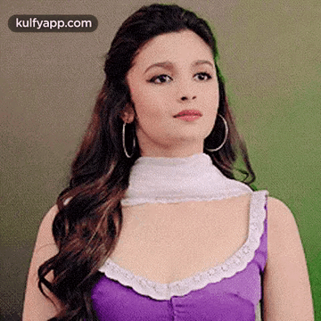 A Woman Wearing A Purple Top And Hoop Earrings Is Standing In Front Of A Green Wall . GIF