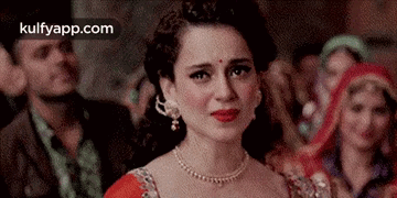 A Woman Wearing A Red Dress And A Necklace Is Crying In Front Of A Crowd . GIF