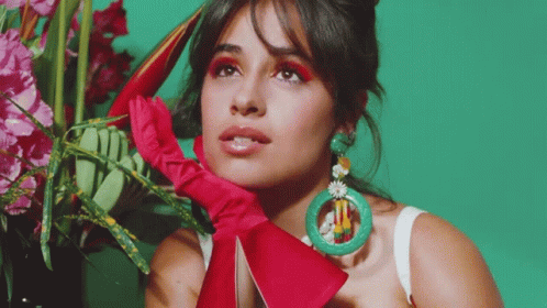 A Woman Wearing A Red Glove And Green Earrings Is Standing In Front Of A Vase Of Flowers GIF