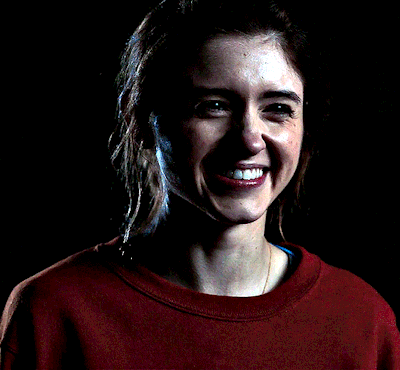 A Woman Wearing A Red Sweatshirt With A Pocket On The Front Smiles In The Dark GIF