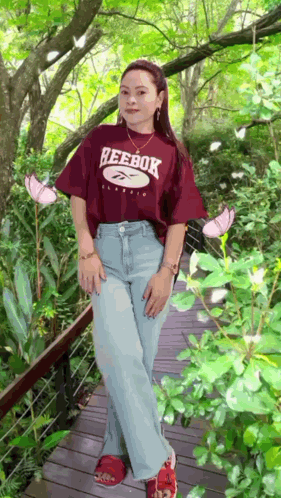 A Woman Wearing A Reebok T-Shirt Is Standing On A Wooden Bridge GIF