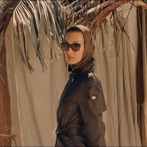 A Woman Wearing A Scarf And Sunglasses Stands In Front Of A Palm Tree GIF
