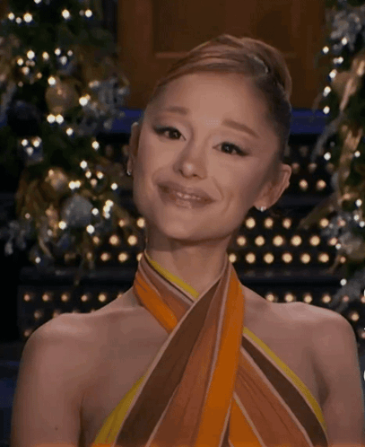 A Woman Wearing A Scarf Around Her Neck Is Smiling In Front Of A Christmas Tree . GIF