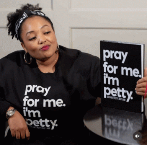 A Woman Wearing A Shirt That Says Pray For Me I 'M Petty Is Holding A Book GIF