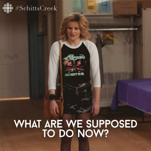 A Woman Wearing A Shirt That Says " What Are We Supposed To Do Now " Stands In A Room GIF