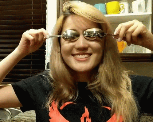 A Woman Wearing A Shirt With A Phoenix On It Is Holding A Spoon To Her Eye GIF