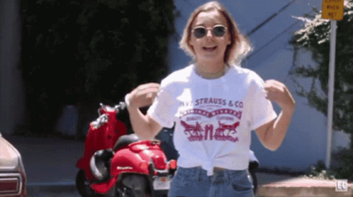 A Woman Wearing A Strauss & Co T-Shirt Is Dancing In Front Of A Red Scooter GIF