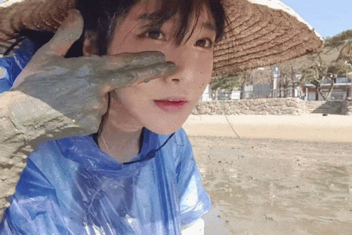 a woman wearing a straw hat and a blue raincoat has a hand covered in mud on her face