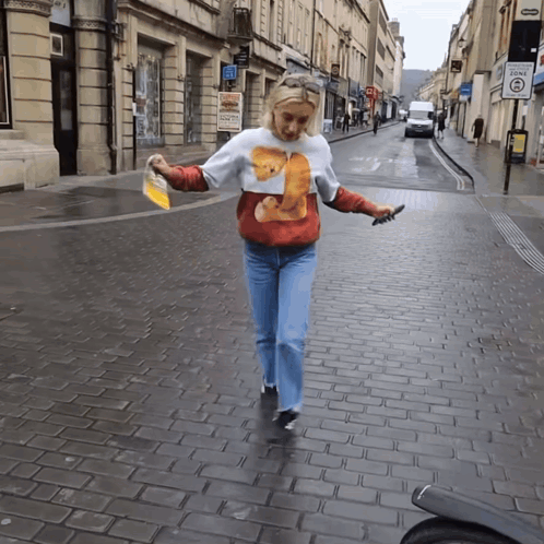 A Woman Wearing A Sweater With A Bear On It Is Walking Down A Brick Street GIF