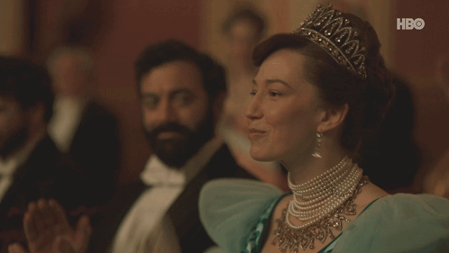 A Woman Wearing A Tiara And Pearls Sits In Front Of A Hbo Sign GIF