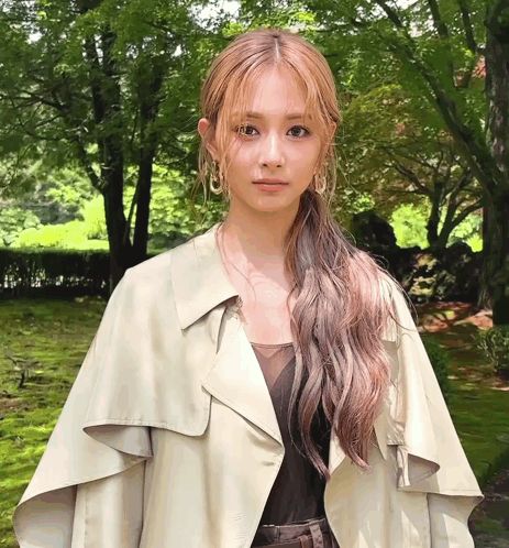 A Woman Wearing A Trench Coat And Earrings Is Standing In A Park GIF