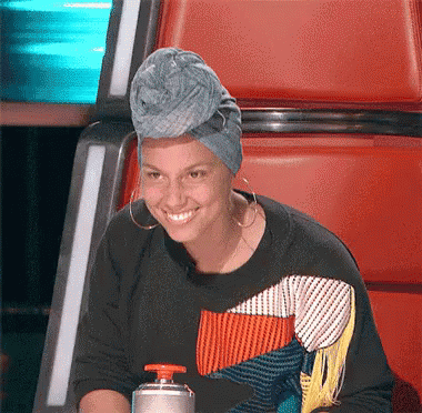 A Woman Wearing A Turban And Earrings Smiles While Sitting In A Chair GIF