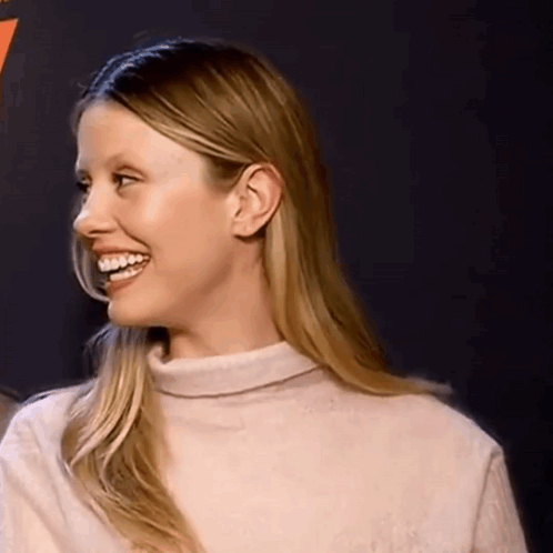 A Woman Wearing A Turtleneck Sweater Is Smiling And Looking To The Side . GIF