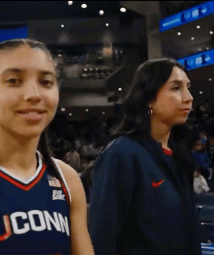 A Woman Wearing A Uconn Jersey Stands Next To A Woman Wearing A Nike Jacket GIF