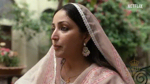 A Woman Wearing A Veil And Earrings Is Sitting On A Bench . GIF