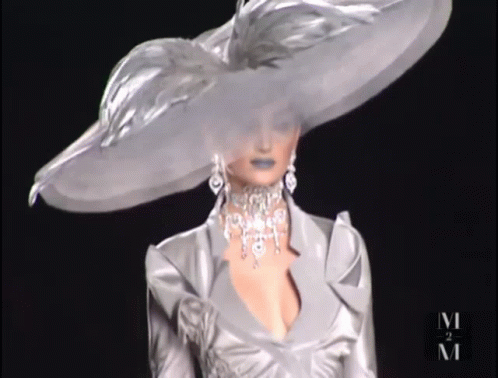 A Woman Wearing A Very Large Hat And A Necklace With The Letter M On The Bottom GIF