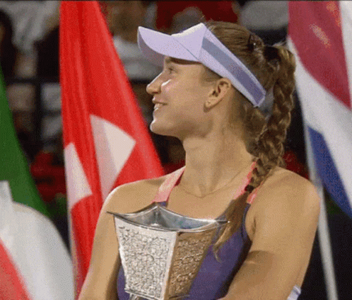 A Woman Wearing A Visor Holds A Trophy In Front Of Flags GIF