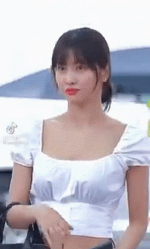A Woman Wearing A White Crop Top Is Standing In Front Of A Car . GIF