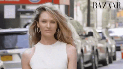 A Woman Wearing A White Dress And Hoop Earrings Is Standing In Front Of A Row Of Cars In A City Street . GIF