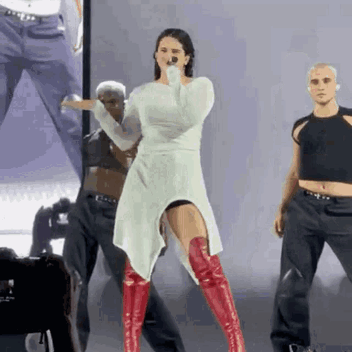 A Woman Wearing A White Dress And Red Boots Is Singing Into A Microphone . GIF