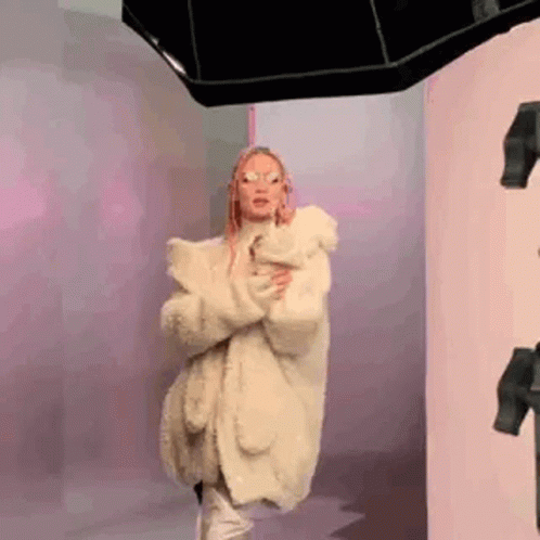 A Woman Wearing A White Fur Coat And Sunglasses Is Standing In Front Of A Mirror . GIF