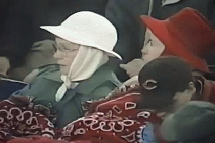 A Woman Wearing A White Hat And A Scarf Is Sitting In A Crowd Of People . GIF