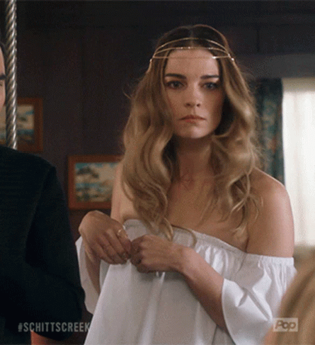 A Woman Wearing A White Off The Shoulder Top Is Standing Next To A Man With #Schittscreek On The Bottom Right GIF