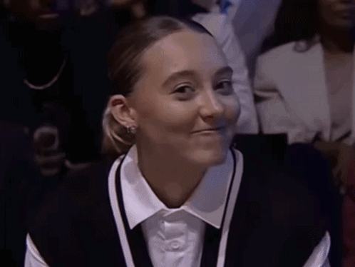 A Woman Wearing A White Shirt And A Black Vest Smiles While Sitting In A Crowd GIF