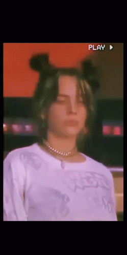 A Woman Wearing A White Shirt And A Necklace Looks At The Camera With A Play Button In The Corner GIF