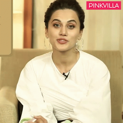 A Woman Wearing A White Shirt And Hoop Earrings Sits In Front Of A Pinkvilla Logo GIF